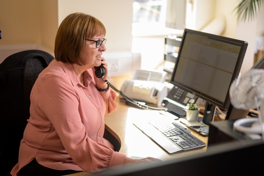 A woman is in her workplace sat at a desk in front of the computer whilst picking up a telephone call.