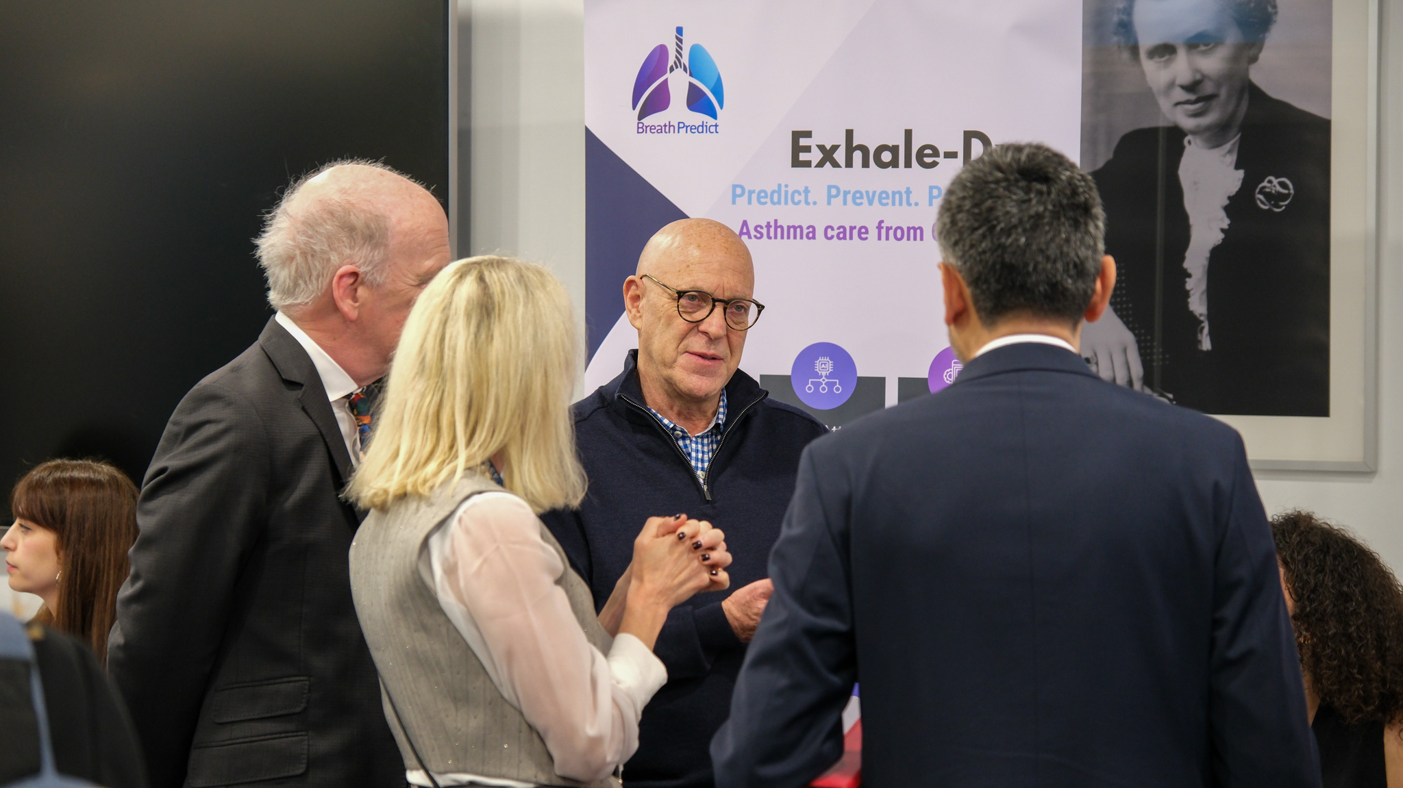 Four people stand in a huddle, talking. In the background, a banner displays the text 'Exhale-D... Predict. Prevent.'