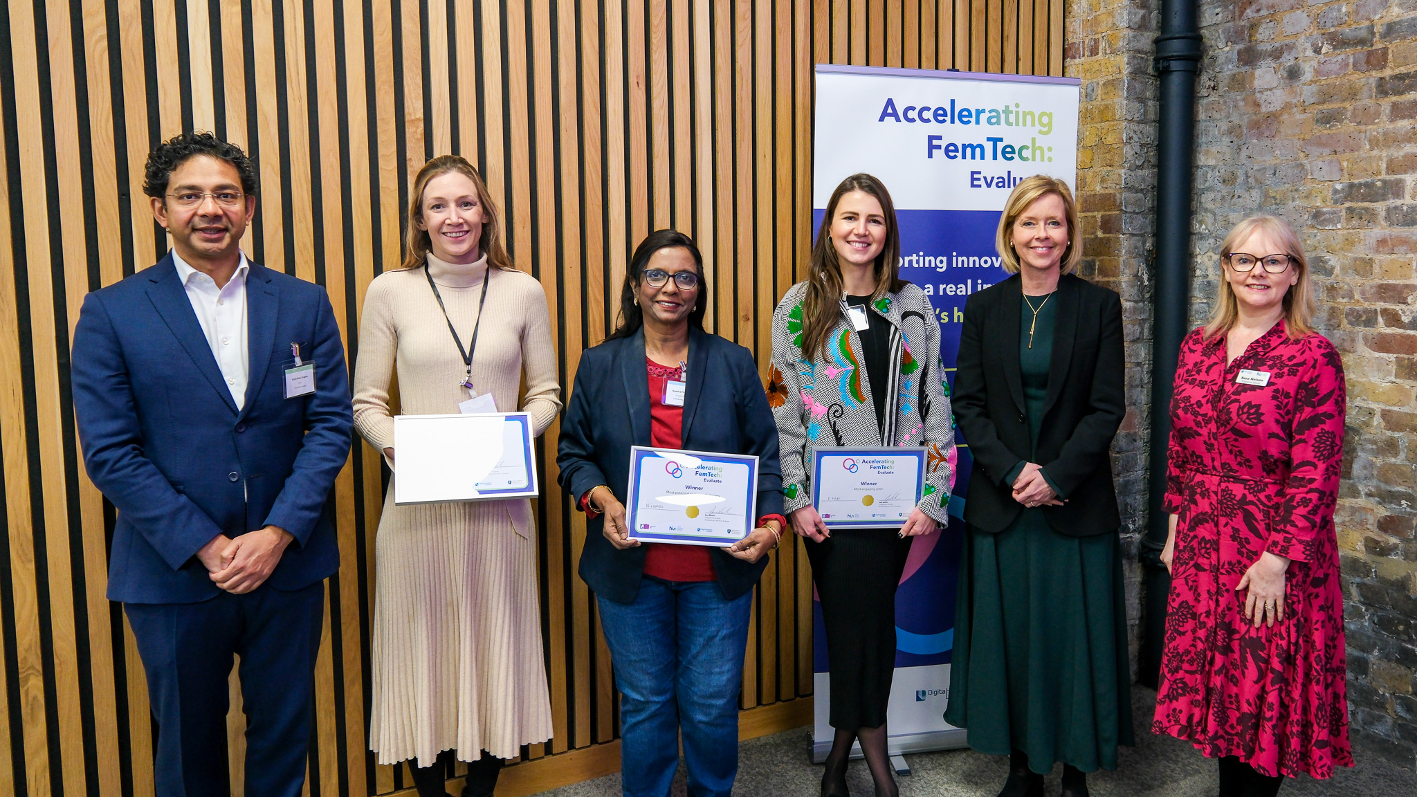 Six people stand in a row. The middle three people hold frames with certificates. In the background, there is a wooden wall and a thin banner that displays the text 'Accelerating FemTech: Evaluate'